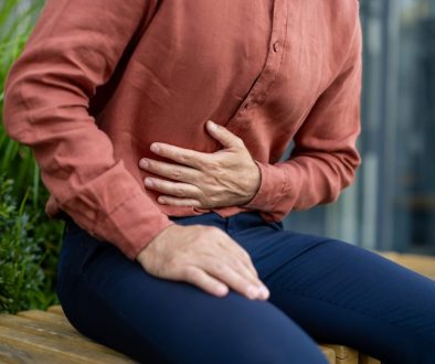 Man in casual attire sitting outdoors holding stomach showing symptoms of discomfort possibly indicating pain or digestive issues wearing brown shirt and dark pants isolated in urban setting mengenal gerd