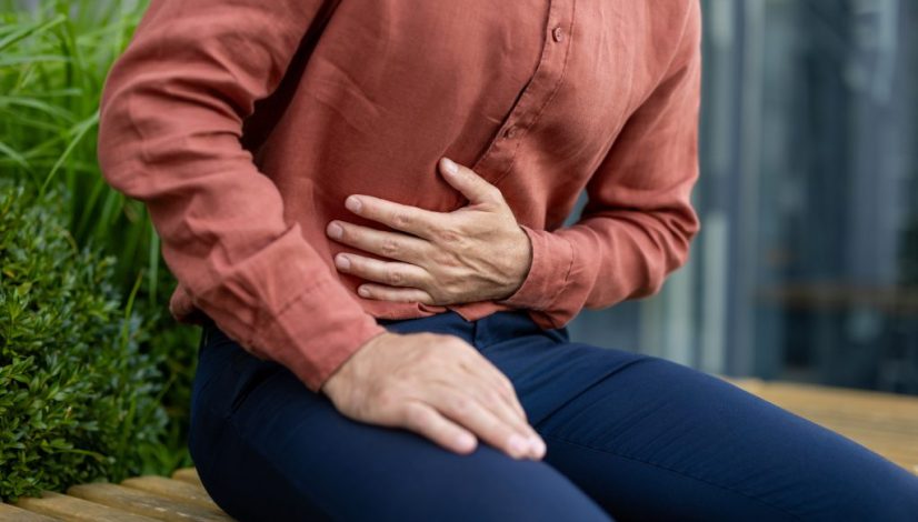 Man in casual attire sitting outdoors holding stomach showing symptoms of discomfort possibly indicating pain or digestive issues wearing brown shirt and dark pants isolated in urban setting mengenal gerd