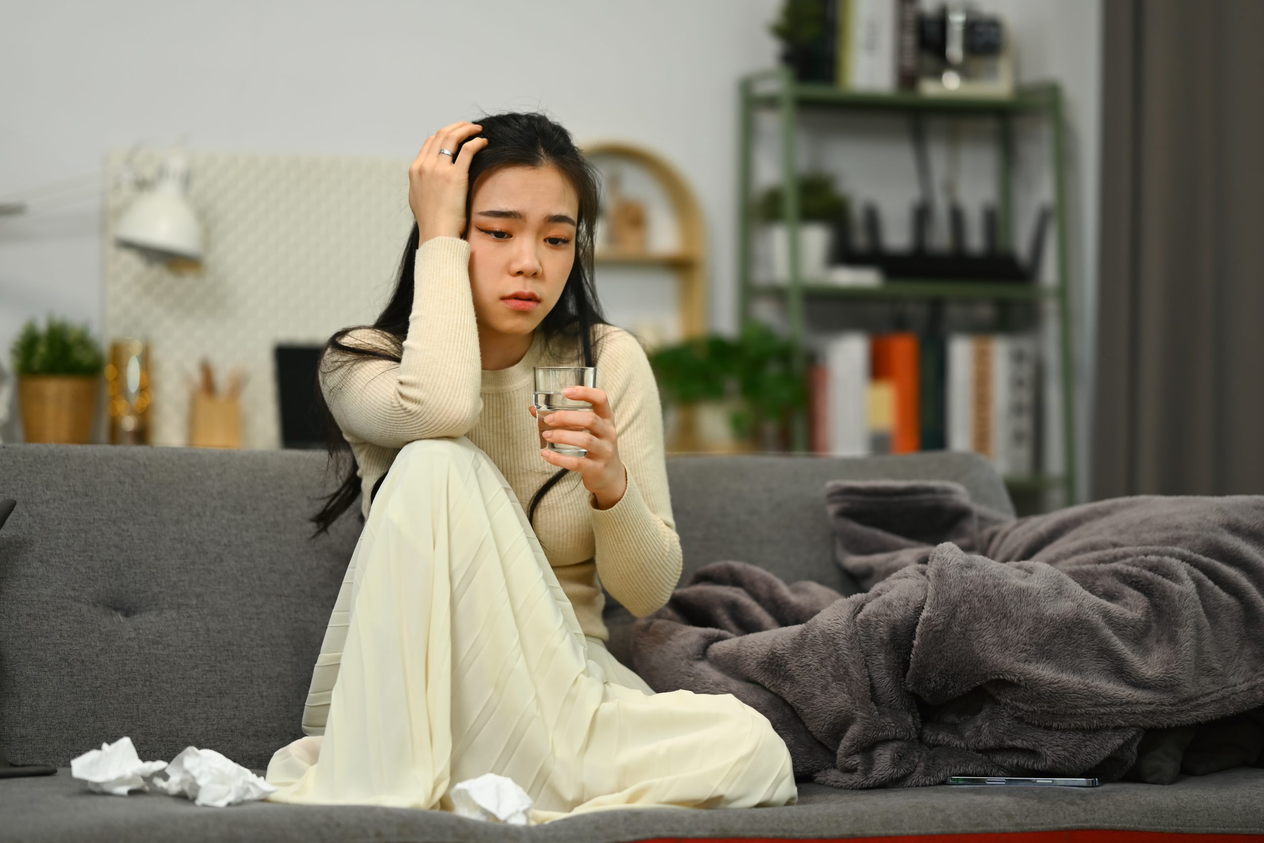 Sick woman sitting on sofa at home with glass of water holding her head, suffering from cold