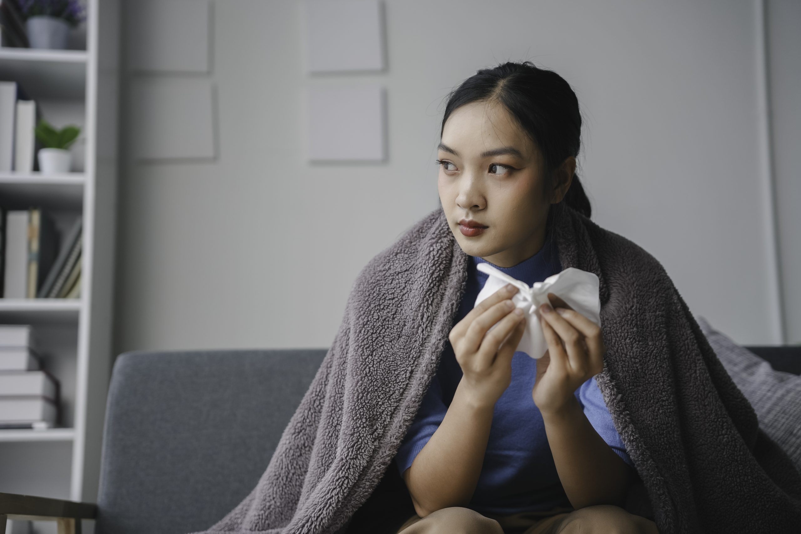 Young woman is sitting on a sofa, covered with a blanket and holding a tissue, feeling unwell